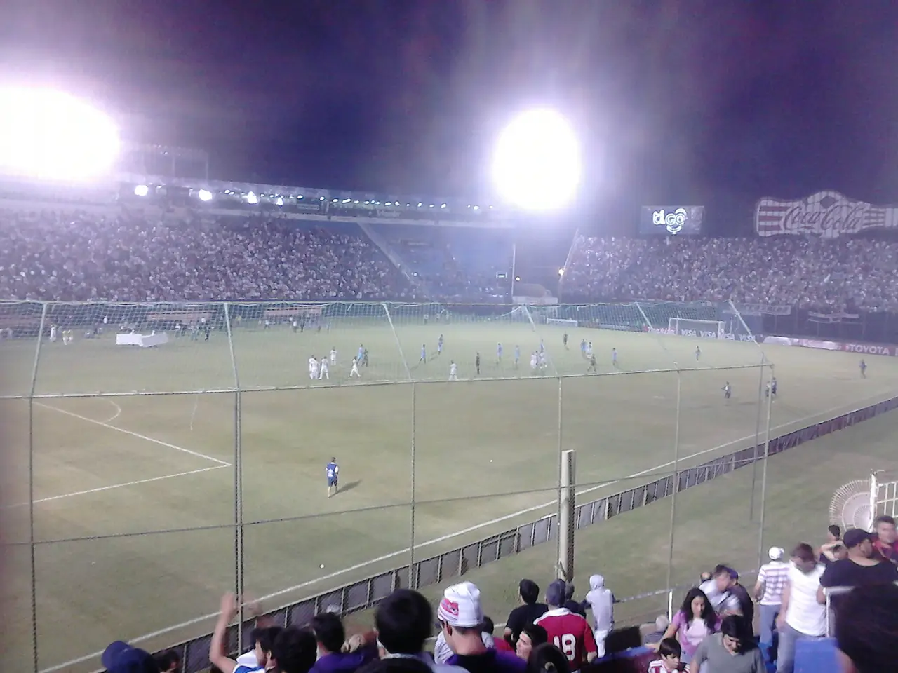 Estadio visto desde la tribuna de noche, con reflectores encendidos y la cancha al fondo.