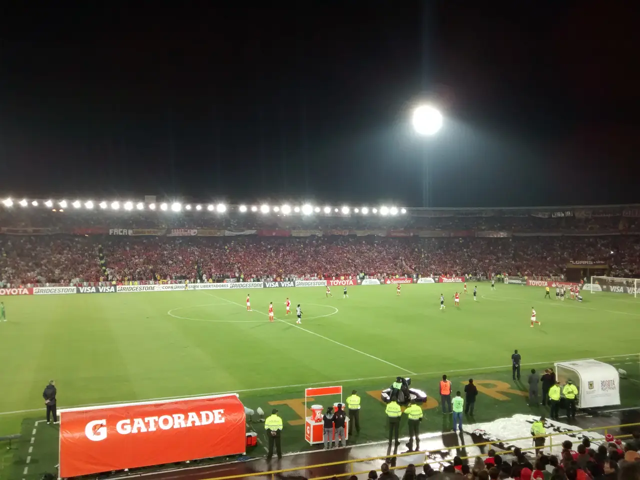 Estadio lleno de noche con reflectores encendidos y hinchada de rojo sobre la cancha.