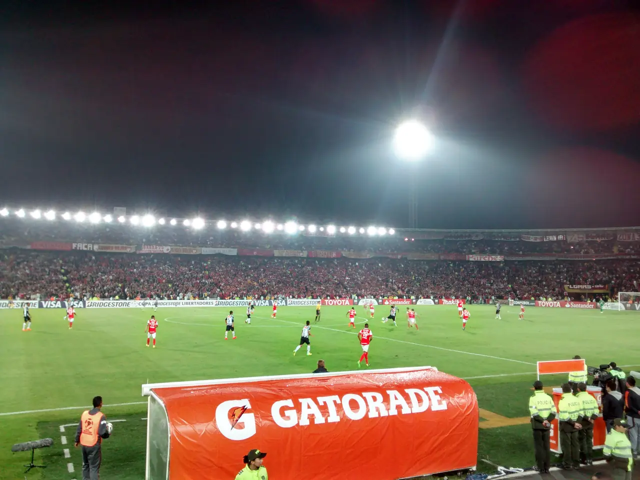 Campo de juego de noche con reflectores, una valla publicitaria roja en primer plano y público al fondo.