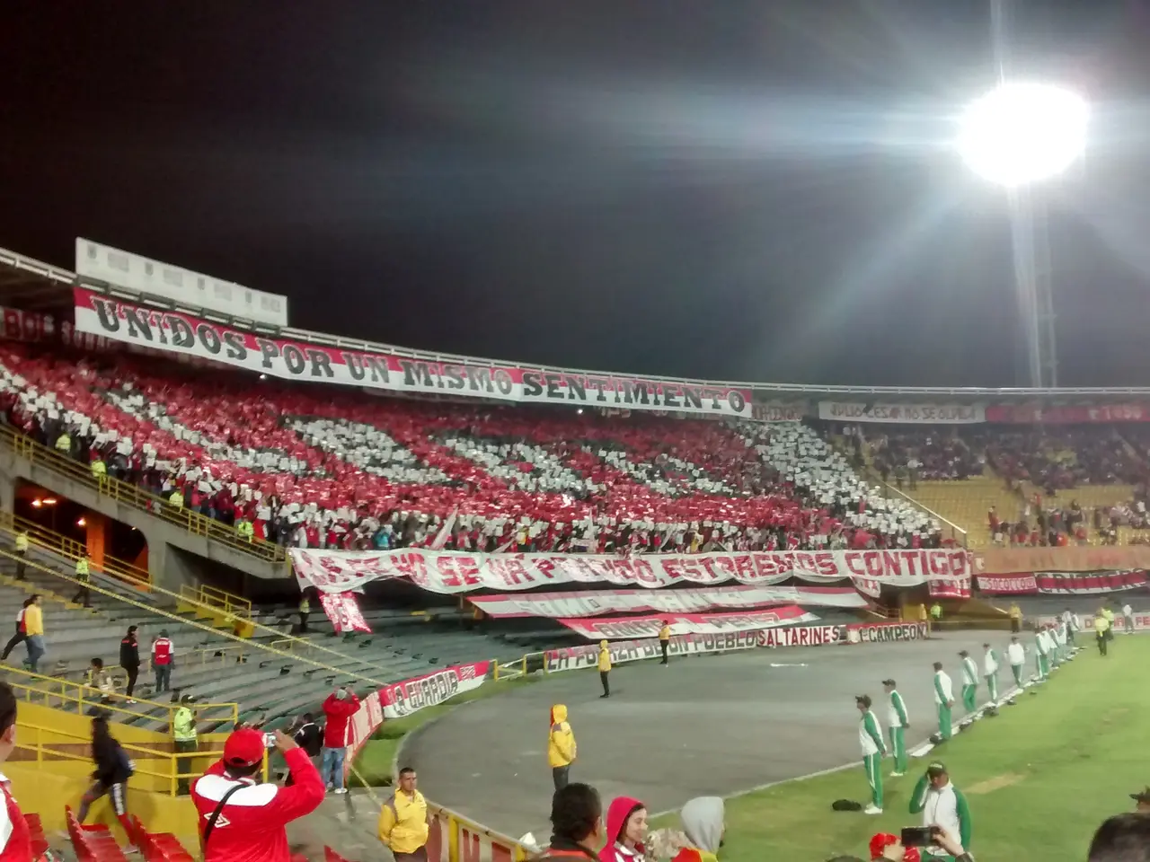 Tribuna popular con banderas y mosaico de hinchada vista desde el costado de la cancha, de noche.